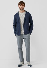 Marineblauer Strickcardigan mit Schalkragen, zwei Vorderentaschen, über einem leicht strukturierten Pullover getragen, kombiniert mit hellgrauen Hosen und Turnschuhen.