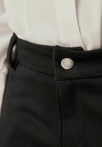 Mustad püksid, millel on sile kangas, nähtav vööloop ja hõbedane metallnupp, millel on graveeritud 'Clarks'.