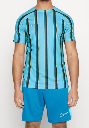 Homme portant un maillot de sport Nike bleu à rayures et un short Nike bleu uni, debout devant un fond blanc.