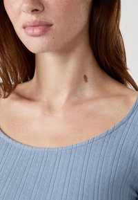 Jeune femme avec de légères taches de rousseur, portant un haut côtelé bleu clair, montrant une naissance brune sur le côté droit de son cou.