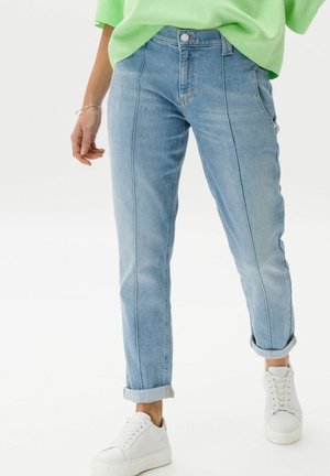 Personne portant un jean bleu clair retroussé, des baskets blanches, un haut vert citron et un bracelet argenté, debout sur fond blanc.