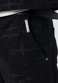 Pantaloni neri con un motivo sottile di stelle e il testo "PXP", completi di un'etichetta argento con la dicitura "PROJECT X PARIS" e un dettaglio del bottone argentato.
