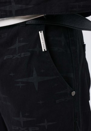 Pantalones negros con un sutil patrón de estrellas y texto "PXP", con una etiqueta plateada que dice "PROJECT X PARIS" y un detalle de botón plateado.
