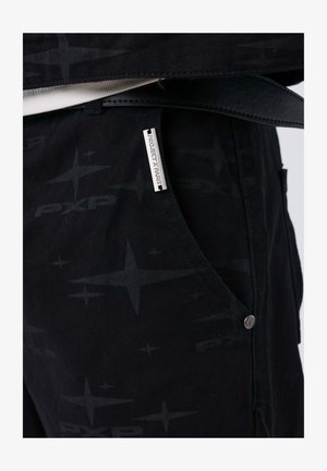Pantaloni neri con un motivo sottile di stelle e il testo "PXP", completi di un'etichetta argento con la dicitura "PROJECT X PARIS" e un dettaglio del bottone argentato.