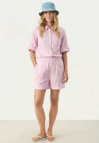 Pink linen short-sleeve shirt with a pocket, paired with matching pink shorts featuring an elastic waistband. Blue bucket hat and sandals.