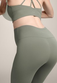 Olivno zelena športna modrček in hlače set, narejen iz gladkega, raztegljivega materiala. Vključuje križno naramnice in subtilno blagovno znamko na pasu.