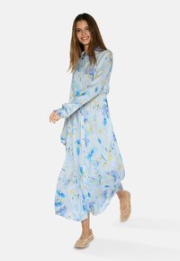 Rochie lungă de culoare albastru deschis, cu print floral în nuanțe de albastru și galben. Material moale, cu front cu butoane și maneci lungi. Asortată cu pantofi bej, plat.