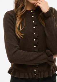 Brūns vilnas kardigans ar apaļu kakla izgriezumu, pogu aizdari, rumbu priekšpusi un piedurknes, kā arī pieguļošu apakšmalu. Mīksta tekstūra ar vieglu izskatu.