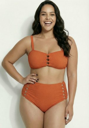 Kvinde med langt mørkt hår smiler iført tekstureret orange todelt badedragt med udskæringsdetaljer på toppen og høj talje på bunden.