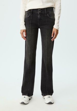 Persoon in zwarte wijde jeans met zichtbare voornaad, witte lange mouwtop en witte sneakers tegen een effen achtergrond.