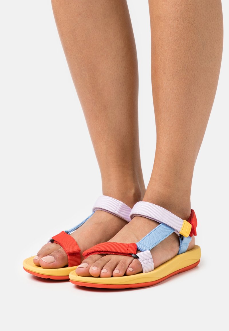 Pieds portant des sandales colorées avec des sangles rouges, bleues et blanches, des semelles jaunes et une boucle rouge et jaune sur un fond blanc uni.