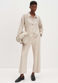 Beige op maat gemaakte jas met knopen en opgestikte zakken, gecombineerd met een broek met wijde pijpen. Het model houdt een bijpassende beige tas vast en draagt zwarte loafers.