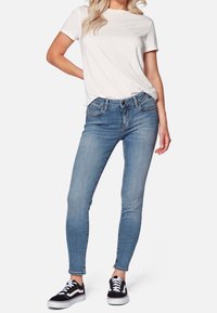 Jeans ajustados de mezclilla azul con un lavado claro, diseño de cinco bolsillos y cintura de altura media, combinados con una camiseta blanca básica y zapatillas negras.