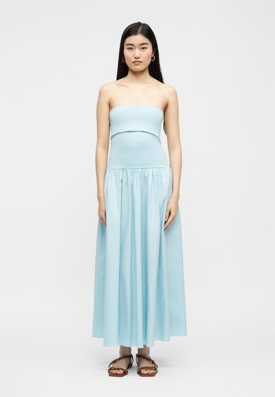 Γυναίκα που στέκεται φορώντας ένα αμάνικο, γαλάζιο, μακρύ φόρεμα μέχρι το πάτωμα και καφέ επίπεδα σανδάλια, μπροστά σε απλό λευκό φόντο.