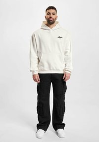 Cremefarvet hoodie med en frontlomme og sort logo, parret med sorte cargo-bukser med flere lommer og hvide sneakers.