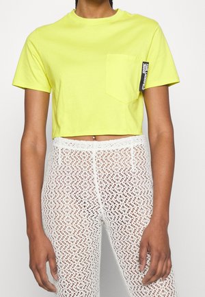 Person iført neongul cropped t-shirt med brystlomme og hvide high-waisted blondebukser, der viser en navle piercing.