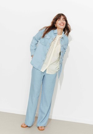 Femme souriante, penchée en avant, portant une veste et un pantalon en denim bleu clair avec un chemisier crème et des sandales marron sur fond blanc.