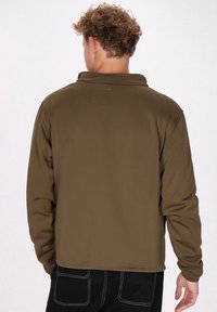 Bruine, fleece-gevoerde pullover met een kraag, voorzien van een rechthoekige patch op de bovenrug en contrasterende stiksels. Lange mouwen.
