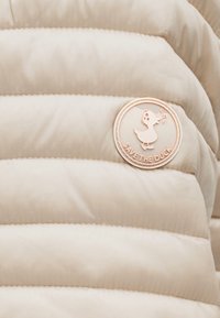 Ύφασμα μπεζ μπουφάν με επένδυση, με στρογγυλή λογότυπη μπαλώμα που δείχνει μια πάπια που τραγουδάει και το κείμενο «SAVE THE DUCK.»