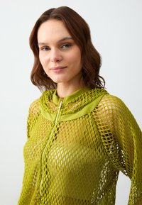 Grüne Netzjacke mit Reißverschluss und Kapuze sowie einem einfarbigen grünen Kragen. Sie zeichnet sich durch ein loses, offenes Webmuster mit auffälligem Reißverschlussdetail aus.