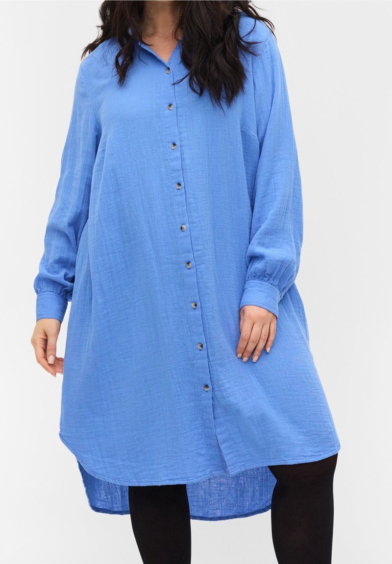 Robe-chemise bleue en tissu texturé, avec des manches longues, un ourlet courbé et des boutons en métal sur le devant.