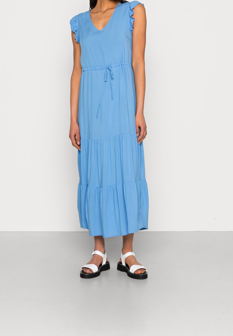 Helles blaues Midikleid mit V-Ausschnitt, Rüschenshuldren und einem gebundenen Taillengürtel. Mit einem gestuften Rock, der bis zur Mitte der Waden reicht. Getragen mit weißen Sandalen.