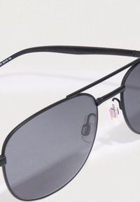 Μαύρο μεταλλικό πλαίσιο αεροπόρων γυαλιών ηλίου με σκούρους γκρίζους φακούς, ακουμπισμένα σε λευκή επιφάνεια, δείχνοντας τα μαξιλαράκια της μύτης και ένα μέρος του βραχίονα.