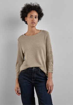 Maglietta a maniche lunghe beige con vestibilità rilassata, scollatura rotonda e leggera lucentezza, abbinata a jeans scuri con tasche frontali.