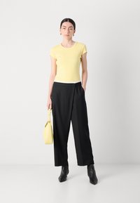 T-shirt jaune ajusté à manches courtes, associé à un pantalon noir ample. Le mannequin tient un petit sac jaune et porte des bottes noires à la cheville.