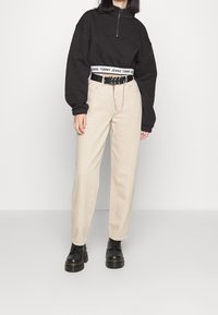 Černá mikina na zip s bílým logem na pase, spárovaná se světle béžovými rovnými kalhotami a černými masivními šněrovacími botami.