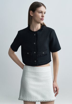 Donna che indossa una giacca corta nera a maniche corte con bottoni e una minigonna bianca testurizzata con orlo decorato con perline, in piedi davanti a uno sfondo grigio.