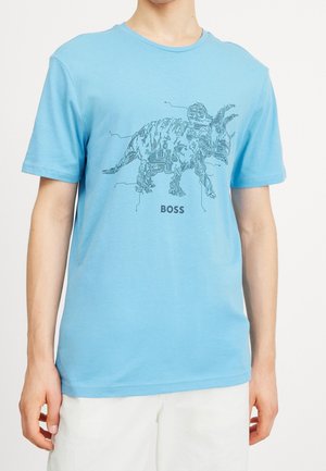 Persoon die een lichtblauw T-shirt draagt met een print van een triceratops in printplaatstijl en het woord "BOSS" eronder, gecombineerd met witte shorts.