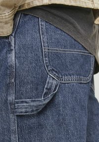 Pantaloni cargo din denim de culoare albastru închis, cu un buzunar lateral cu clapetă, cusături vizibile și o textură ușor decolorată.