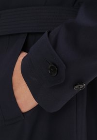 Granatowy mankiet płaszcza przeciwdeszczowego o gładkiej fakturze, z dwoma czarnymi guzikami i przeszyciem nad mankietem, zapewniający pewne dopasowanie.