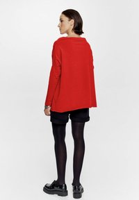 Czerwony, oversize sweter z długimi rękawami, zestawiony z czarnymi szortami. Modelka nosi czarne rajstopy i masywne czarne buty, widoczna z tyłu.