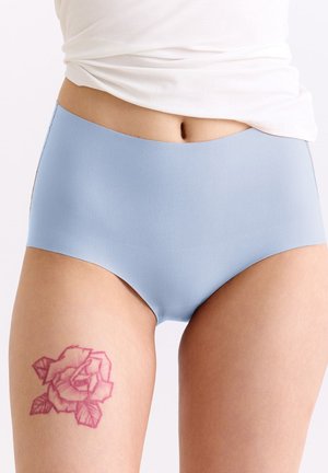 Nahaufnahme einer Person, die nahtlose Unterwäsche in Hellblau und ein weißes Oberteil trägt, mit einem pinken Rosen-Tattoo am oberen Oberschenkel.