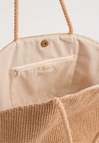 Béžová tkaná taška s pletenými provázkovými uchy. Obsahuje vnitřní zipovou kapsu a zapínání na cvak, má hladkou texturu a jednoduchý design.