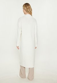 Cardigan alb, tricotat, până la genunchi, cu o textură côtată, având un guler lat și mâneci lungi, asortat cu pantaloni bej.