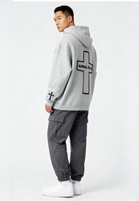 Felpa grigia con cappuccio e testo nero "KARMA KILLS" e croce sul retro; abbinata a pantaloni cargo grigi scuri e sneakers bianche.
