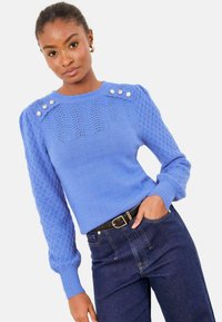 Jasnoniebieski sweter z dzianiny z fakturowanymi rękawami i trzema ozdobnymi guzikami na ramieniu. W połączeniu z ciemnoniebieskimi dżinsami i paskiem.