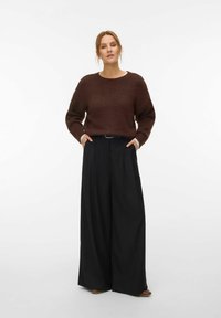 Femme debout, les mains dans les poches, portant un pull en maille marron et un pantalon large noir, sur fond blanc uni.