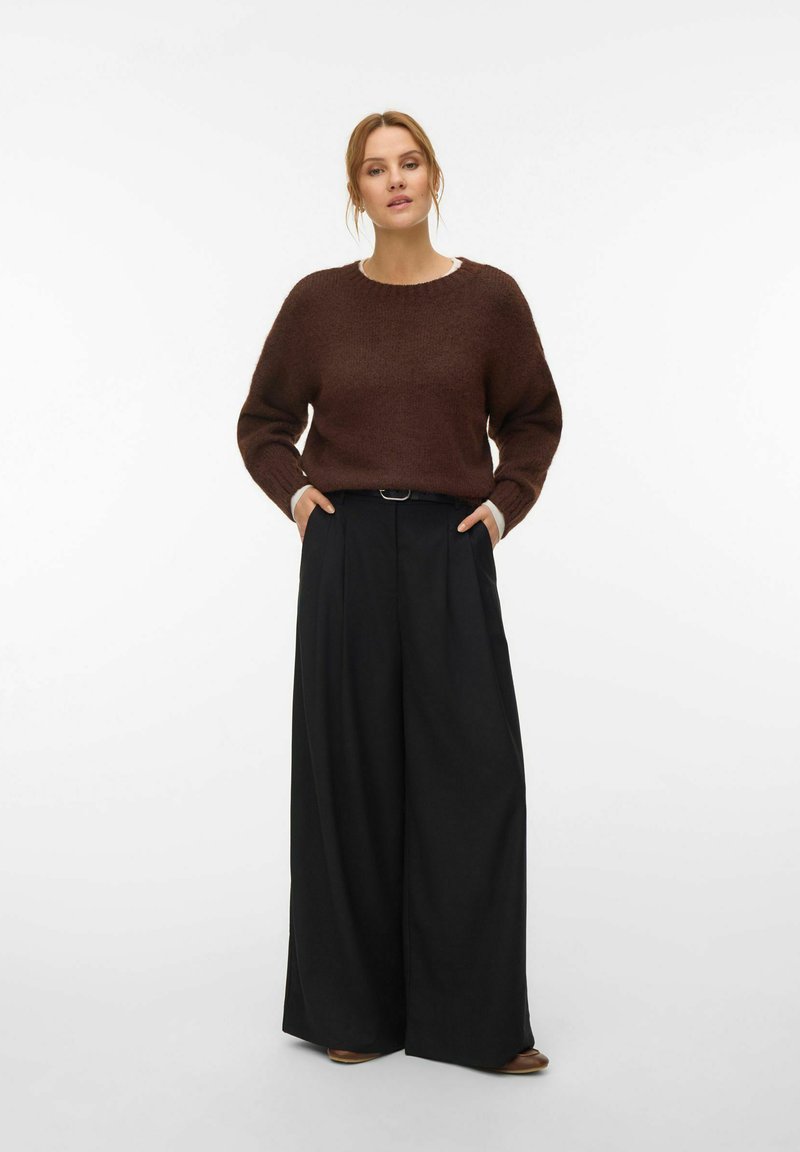 Femme debout, les mains dans les poches, portant un pull en maille marron et un pantalon large noir, sur fond blanc uni.