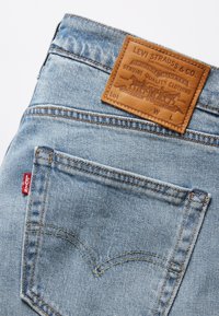 Jeans de mezclilla azul claro con una etiqueta de marca de cuero en la parte posterior. Presentan un parche rojo y costuras amarillas en contraste. Diseño estándar de cinco bolsillos.