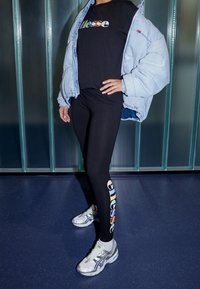 Czarne legginsy z logo "ellesse" w pastelowych kolorach po boku, w połączeniu z jasnoniebieską kurtką puchową i srebrnymi butami sportowymi.