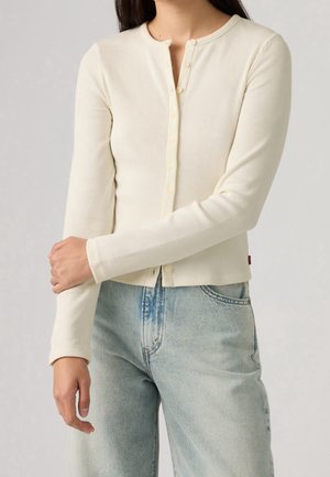 Femme portant un cardigan côtelé crème boutonné et un jean taille haute bleu clair, debout avec un bras croisé sur l'autre.