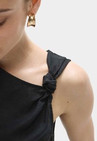 Top nero senza maniche con dettaglio spallina annodata. Tessuto liscio con design aderente. Orecchino in metallo dorato che contrasta con il tessuto.