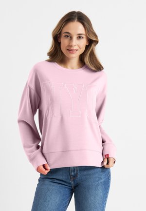 Roze sweatshirt met "NYC" in witte letters, relaxed fit, geribbelde cuffs en onderkant. Gecombineerd met een blauwe spijkerbroek, gepresenteerd op een neutrale achtergrond.