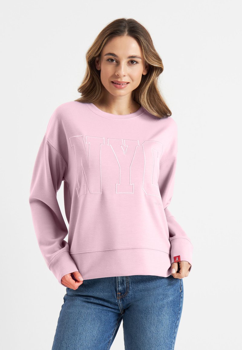 Roze sweatshirt met "NYC" in witte letters, relaxed fit, geribbelde cuffs en onderkant. Gecombineerd met een blauwe spijkerbroek, gepresenteerd op een neutrale achtergrond.