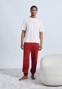 Γκρι βαμβακερό μπλουζάκι με λογότυπο και κόκκινη βαμβακερή φόρμα. Το παντελόνι έχει μανσέτες και πλαϊνές τσέπες. Το μοντέλο στέκεται ξυπόλητο σε ένα ανοιχτό χαλί.