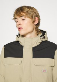 Beige jakke med en svart øverste del, med to framlommer, glidelås-lukking og hette. Teksturert aksenter synlige.